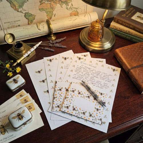 A styled flat-lay of vintage stationery on a wooden desk, featuring scattered letter cards with floral borders, a fountain pen, a small inkwell, a brass lamp, and an old map.