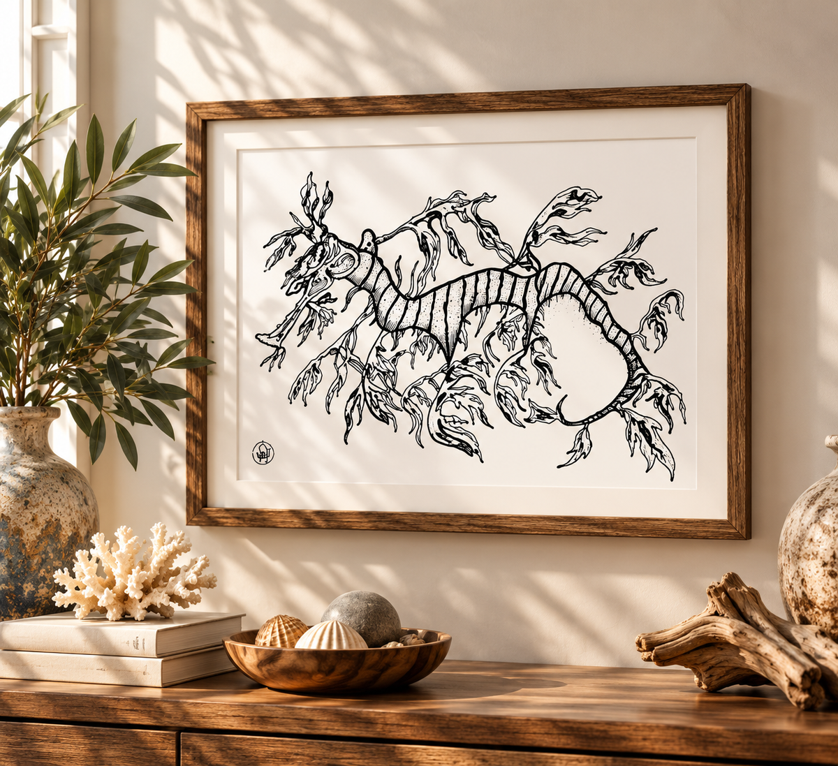 Framed black ink illustration of a sea dragon on a white mat, displayed on a wooden console table in a styled room with plants, shells, and driftwood.