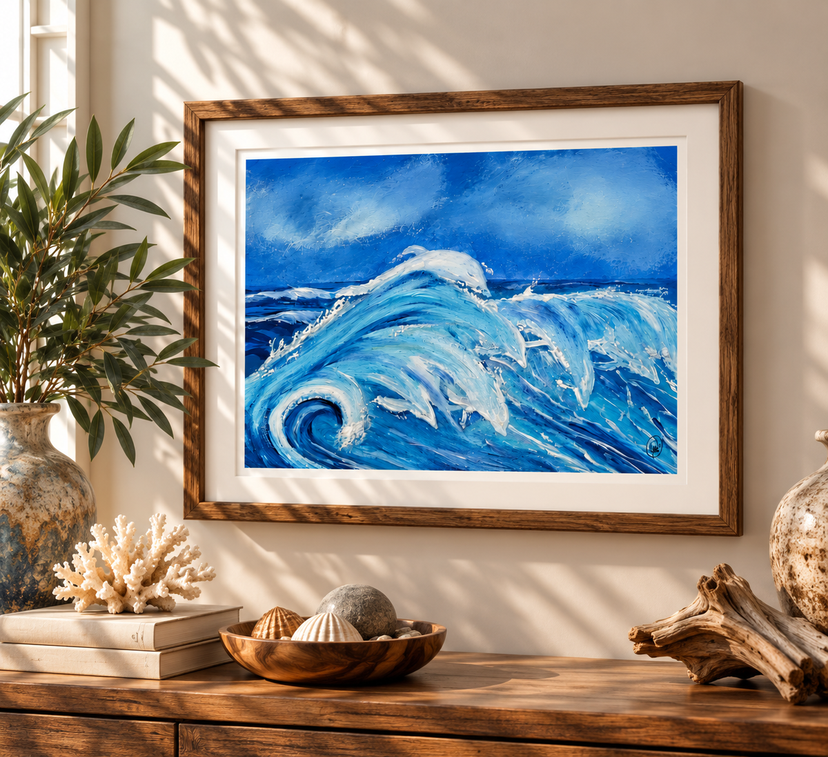 A framed art print depicting a powerful blue ocean wave, styled on a wooden console table with seashells, driftwood, and a potted plant in a sunlit room.