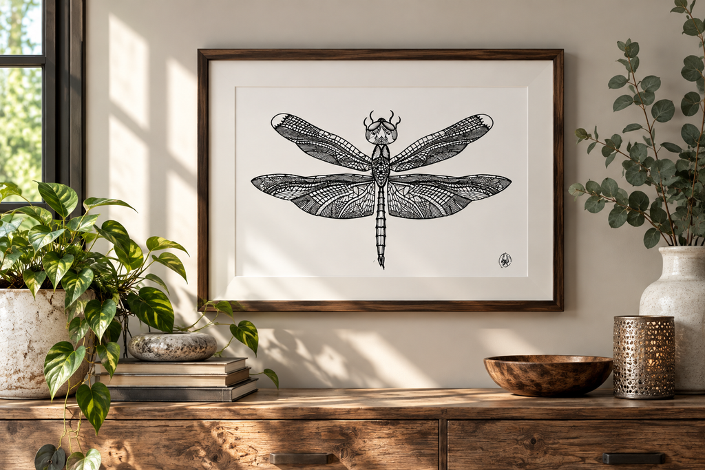 A framed black and white dragonfly art print hanging on a beige wall above a rustic wooden console table, styled with green plants and decorative bowls.