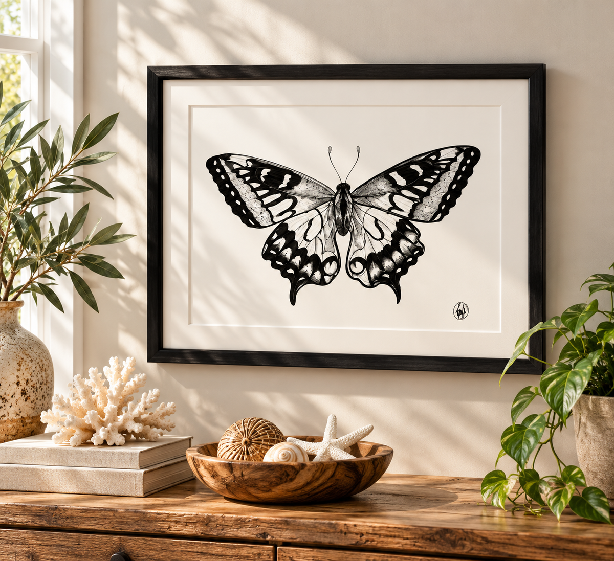 A framed black and white butterfly art print hangs on a beige wall above a wooden console table styled with plants, books, and seashells.
