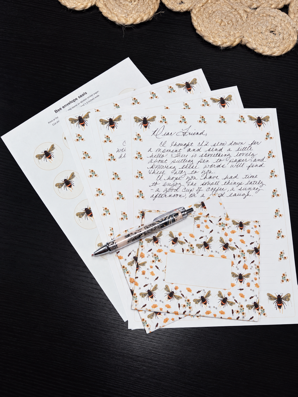 A styled flat-lay of bee-themed stationery, including printed sheets with illustrations of bees, a matching patterned napkin, and a silver pen, arranged on a dark wooden surface with a woven coaster in the background.