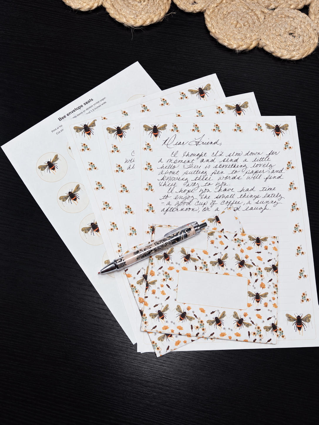 A styled flat-lay of bee-themed stationery, including printed sheets with illustrations of bees, a matching patterned napkin, and a silver pen, arranged on a dark wooden surface with a woven coaster in the background.