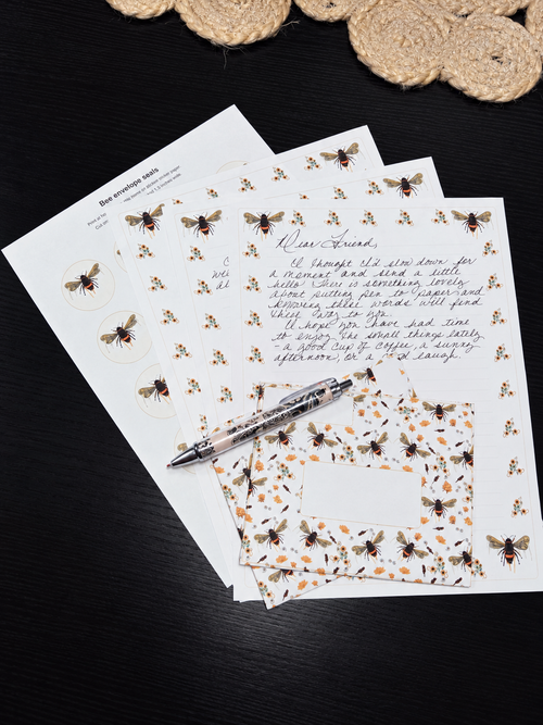 A styled flat-lay of bee-themed stationery, including printed sheets with illustrations of bees, a matching patterned napkin, and a silver pen, arranged on a dark wooden surface with a woven coaster in the background.