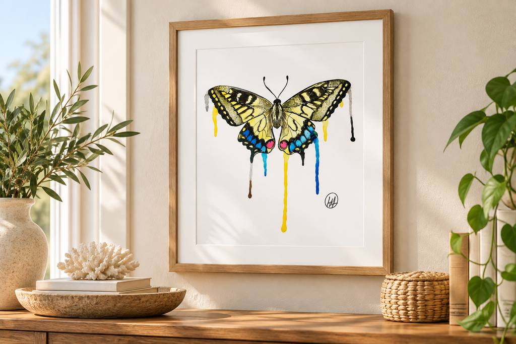 A framed butterfly art print hangs on a beige wall above a wooden console table. The print features a yellow and black butterfly with colorful dripping paint details in blue, pink, yellow, and black, set in a white mat and a light wood frame.