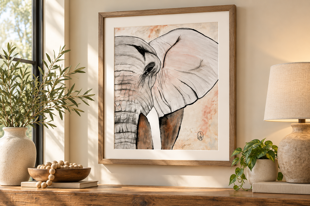A framed art print of an elephant's face, featuring black ink linework and soft pink watercolor washes on a light background, styled on a wooden console table in a warm, sunlit room with a lamp and plants.