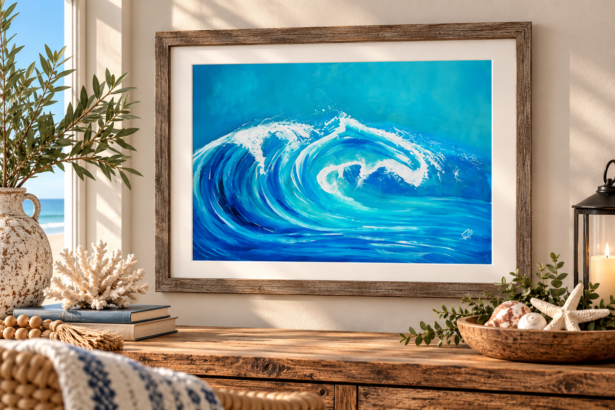 A framed blue ocean wave painting hangs on a beige wall above a rustic wooden console table, styled with a vase, books, coral, and a lantern.