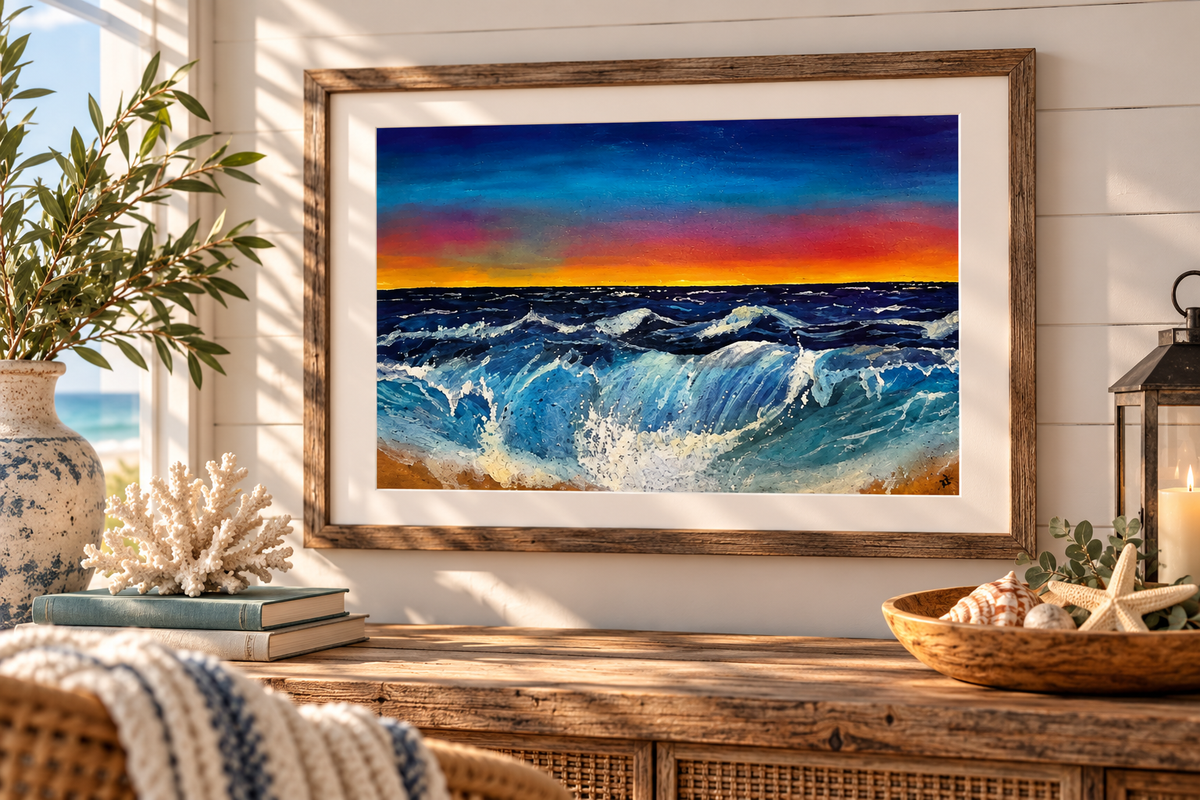 A framed art print depicting a dramatic ocean sunset with deep blue waves and a bright orange-yellow horizon, displayed on a wooden console table in a coastal-themed room with seashells, books, and a plant.